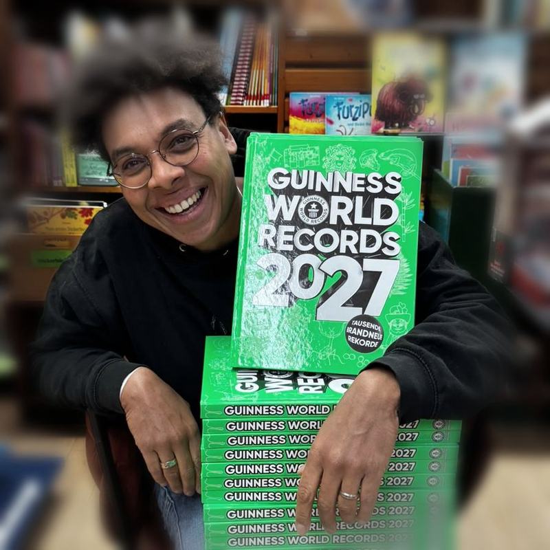 Diana sitzt lachend in ihrer Buchhandlung, die Arme auf einem Stapel Guinness-Bücher gestützt